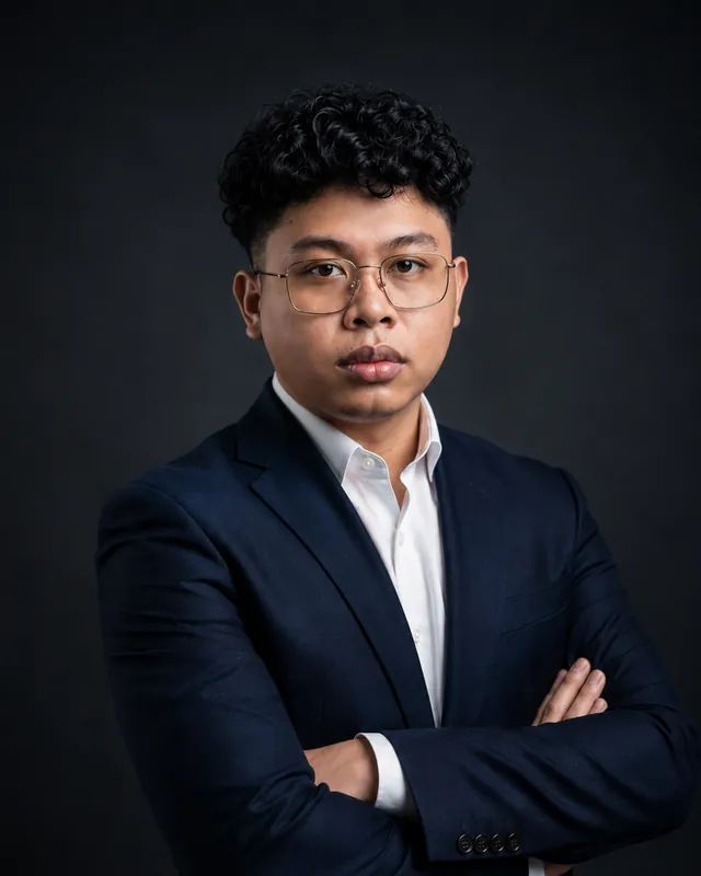Ray Ingotan in a navy suit, arms crossed, against a dark studio background